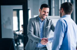 two-smiling-businessmen-shaking-hands-while-standing-office-min