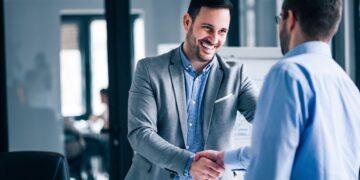 two-smiling-businessmen-shaking-hands-while-standing-office-min